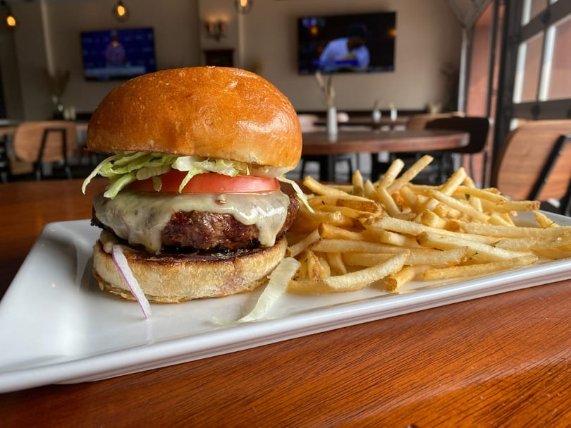 A BARREL HOUSE cheeseburger with lettuce, tomato, and red onion, served with a side of fries.