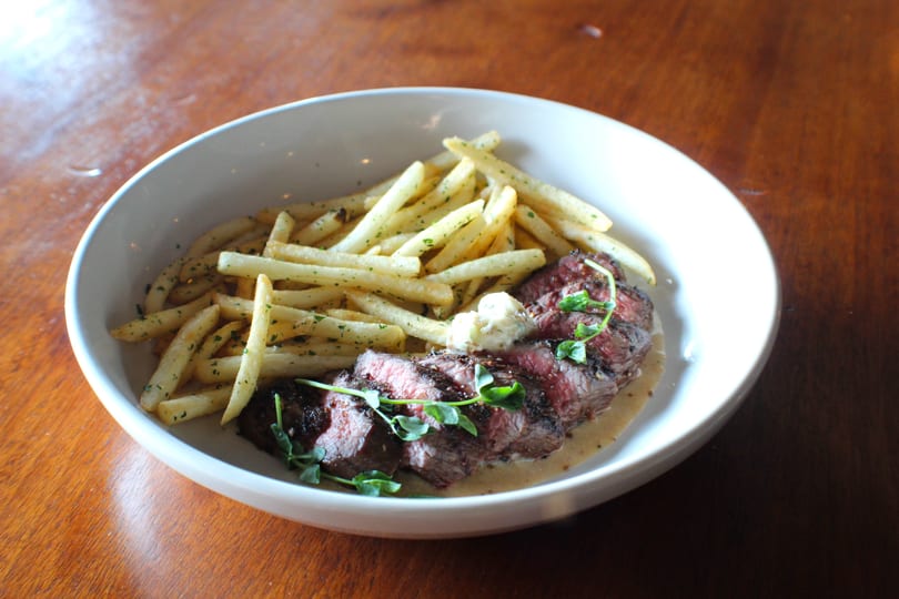 Sliced steak with herb butter, fries, and a creamy sauce in a white bowl on a wooden table.