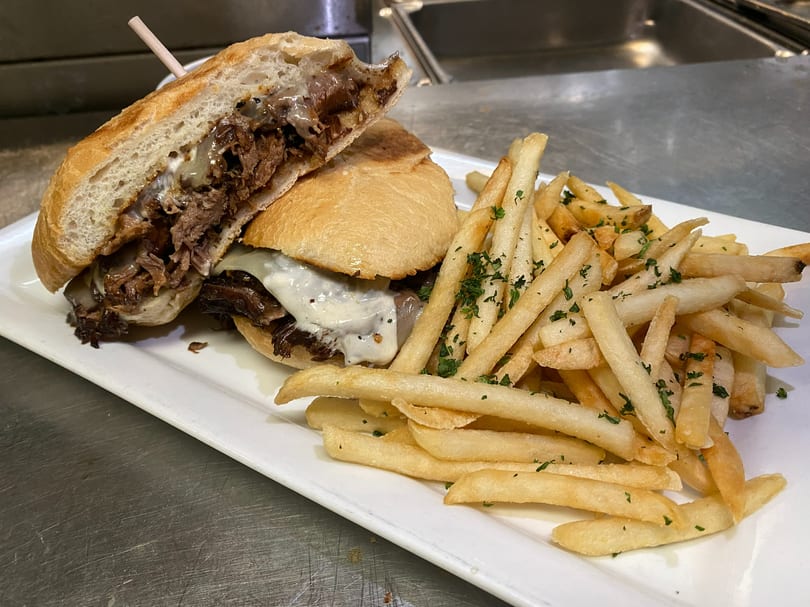 A BARREL HOUSE steak sandwich with melted cheese and caramelized onions, served with golden french fries.