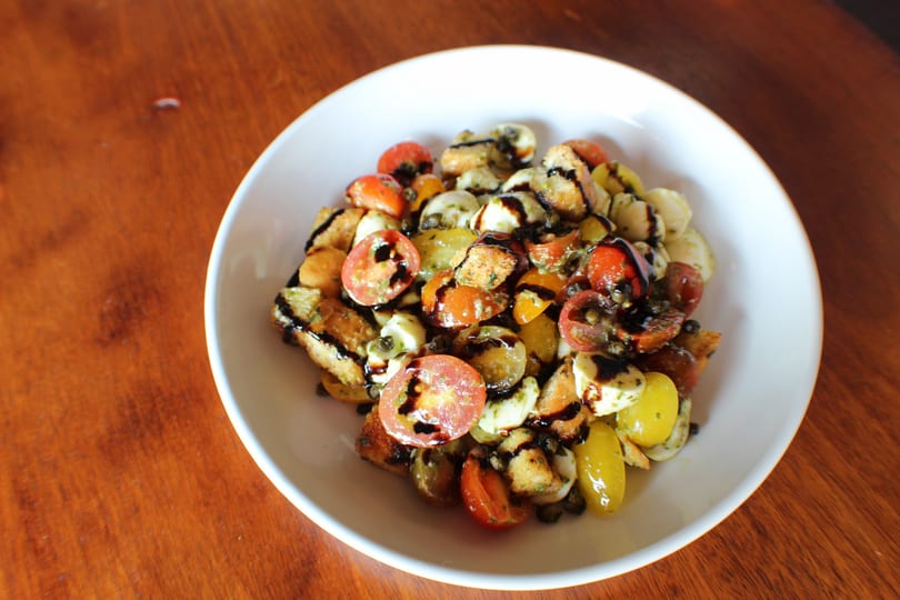A white bowl of panzanella salad with tomatoes, mozzarella, croutons, and balsamic glaze.
