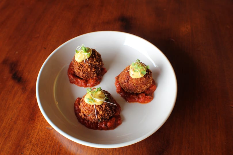 Three crispy croquettes with yellow sauce and microgreens on red marinara in a white bowl.